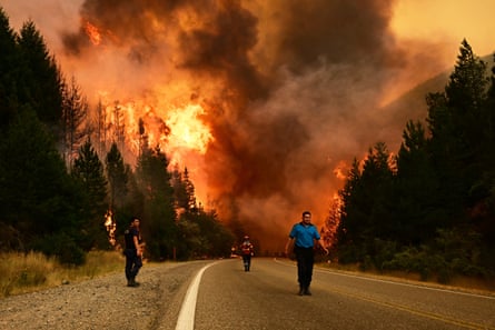 Firefighters walk on a road with towering flames and thick plumes of smoke in the forest behind them.