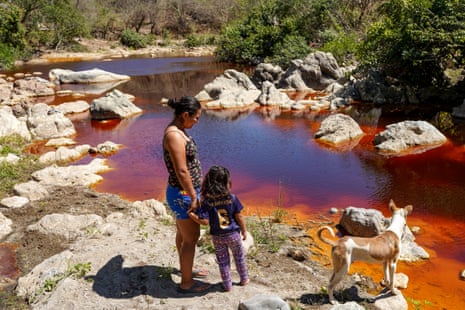 Woman and her daughter on banks of a river coloured red due to substances from mining in Santa Rosa de Lima, El Salvador.