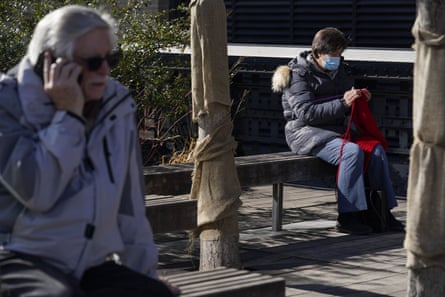 A woman wears a mask while knitting in a park as a man nearby talks on the phone while maskless.
