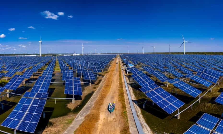 High angle shot of maintenance personnel driving down a dirt road in the middle of a huge array of dark blue solar panels, with numerous wind turbines visible on the horizon against a blue sky