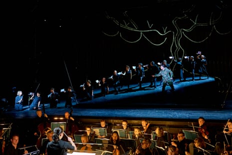 flautist Claire Wickes at the far left of a steeply raked stage, Norman Reinhardt, upstage on the right as Tamino, and row of performers lined up between them half crouching. in the foreground, the ENO orchestra in the pit conducted by Erina Yashima, in The Magic Flute