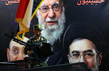 Am man in military fatigues and cap stands behind a mounted gun in front of a poster featuring Ayatollah Ali Khamenei and Ayatollah Mojtaba Khamenei