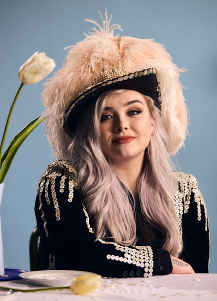 Shannon Crowe in a black cowgirl hat covered in fake white fur as well as her mother-of-pearl pearly outfit, sat next to a table with a white flower on it