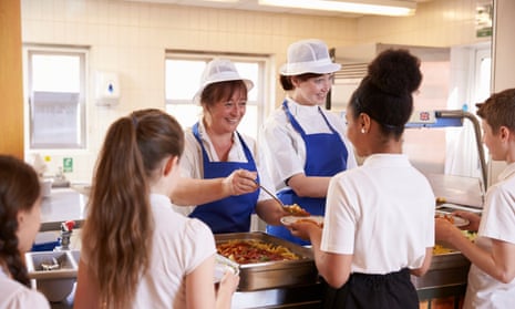 Two women serving kids food in a school cafeteria, back view
