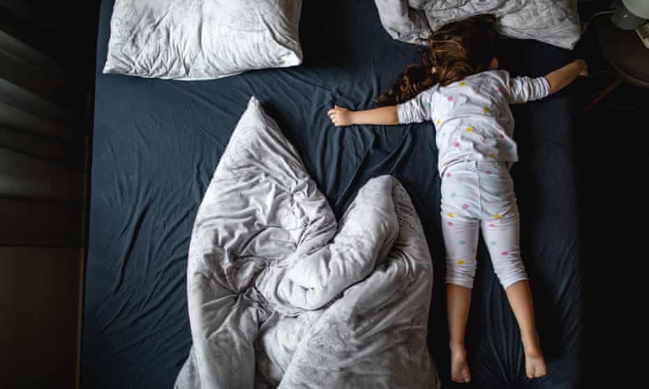 A young girl lies on her stomach on a large bed with her arms and legs stretched out