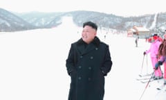 A man wearing a black overcoat stands, smiling, at the bottom of a ski slope