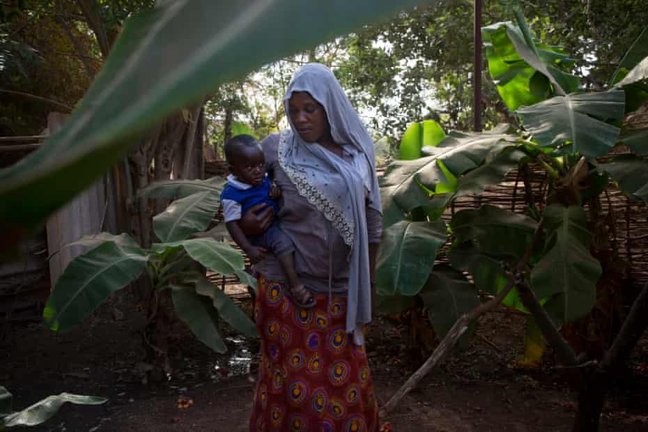 Fatou Ceesay, de 33 años, sostiene a su hijo Ibrahim de 11 meses y camina por el jardín frente a su casa hasta su oficina en el centro de Gambia.