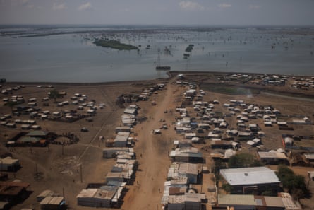 A wide and wiggly dirt road lined with tin shacks leads to a large body of water.