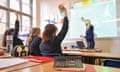 pupils seen from rear with their hands up as teacher points to whiteboard