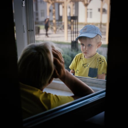 A boy in a yellow T-shirt and blue cap looks in through a window at a person seen from behind looking out