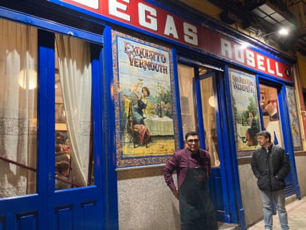 A man in apron standing outside a blue painted bar with red and white signage saying ‘Bodegas Rosell’ and vintage tiled adverts for vermouth and wine