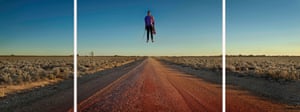 A man floats above a desert road in the middle of the outback holding a violin in one hand and the violin bow in the other.