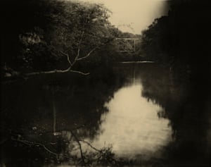 The still water of a river reflects the sepia sky and black trees, with a wooden bridge frame just visible in the distance