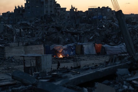 A fire burns in a makeshift shelter, amid scenes of destruction.