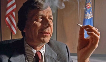 An older man in grey suit smoking with US flags in background