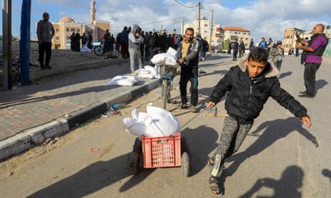 A boy drags bags of flour in a plastic crate along a street followed by a man carrying flour bags on a bike. Behind are other people collecting flour, a mosque and residential buildings.