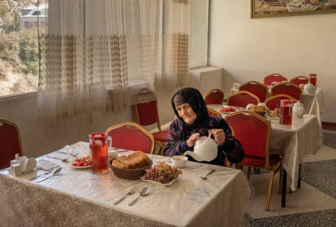 A woman in a headscarf pours tea from a white teapot at a table set with bread and fruit