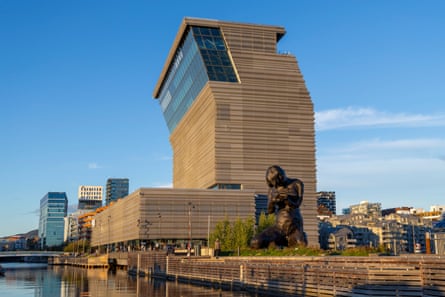 A modern building that slopes on one side, next to the water. A bronze statue is next to the building.