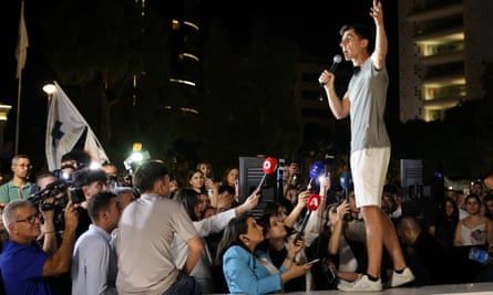 Fidias Panayiotou speaks during celebrations in Nicosia after his election: he is in his 20s and wears a grey T-shirt, white shorts and white trainers; he is standing on a platform in a public square with one arm raised, holding a microphone and addressing a crowd of media reporters and supporters
