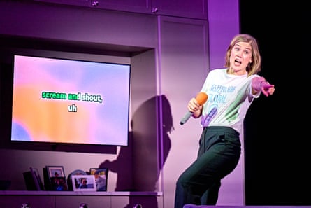 Rosamund Pike singing karaoke on stage, pointing at someone out of sight and holding a mic. Text on the TV screen reads 'scream and shout'