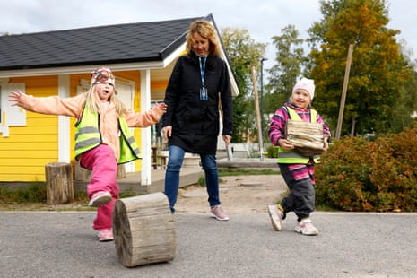 A woman holds the hands of two children wearing hi-vis playing with large wooden blocks