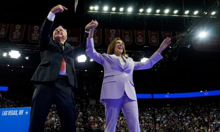 A man (Tim Walz) and woman (Kamala Harris) wearing suits hold hands and they raise their arms in the air at a political rally