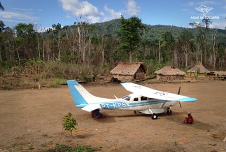 A microlight plane in front of a group of grass huts next to the rainforest