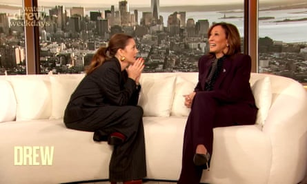 In front of a landscape of Los Angeles skyscrapers, Drew Barrymore, long hair in a ponytail and wearing a suit, leans forward with her hands pressed together as Kamala Harris, laughing and also in a suit, talks with her on a curved sofa