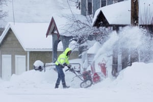 Snow sprays as a man uses a clearing machine next to residential buildings