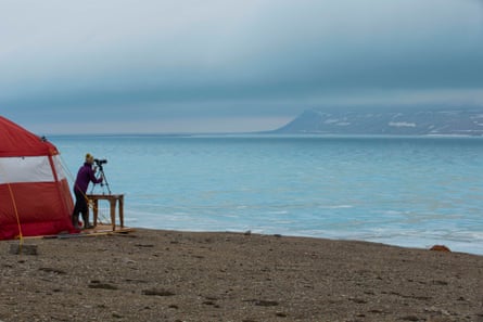 A woman outside a tent on a beach looking through a monocular