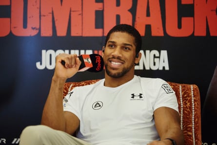 Anthony Joshua in a white T-shirt holds a microphone while sitting in a chair