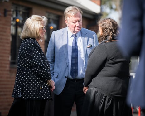 A man in a pale blue jacket stands talking to other guests outside a brick building.
