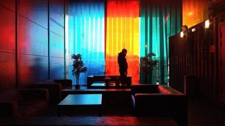 A man standing near some sofas in a moodily lit bar, with brightly coloured transparent panels hanging in its large windows