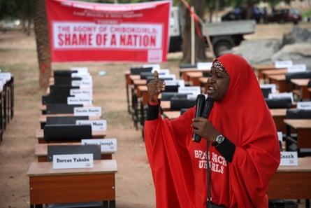 A woman in a red hijab with the slogan Bring Back Our Girls printed on it, speaks into a microphone. In the background are rows of school desks with name cards on them and a banner reading: ‘Agony of Chibok girls. Shame of a nation.’ 