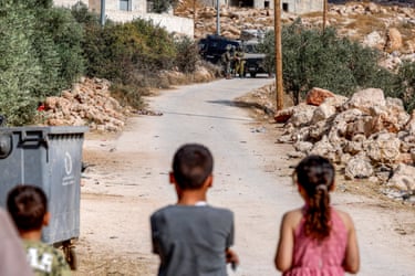 Three young children watch Israeli security forces at the other end of a short track who are standing by their vehicles