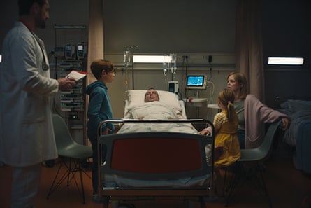 A man lies in a hospital bed surrounded by a worried looking woman, two children and a doctor who is approaching.