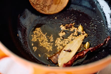 Close up of a saucepan featuring whole cumin, green cardamom, bay leaf and dried chilli in hot oil.