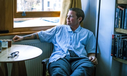 kae tempest seated at a table by a partially opened window in a house with books to one side