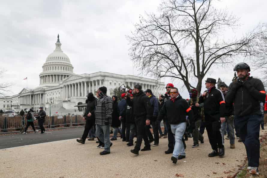 A group of men dressed in black, some with orange armbands and one wearing a helmet, walk by the US Capitol building. 