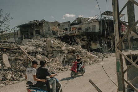 People on mopeds and scooters ride along a road next to piled-up rubble and destroyed buildings