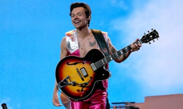 Harry Styles smiling and holding a guitar while wearing a silver and pink metallic waistcoat and matching pink metallic trousers