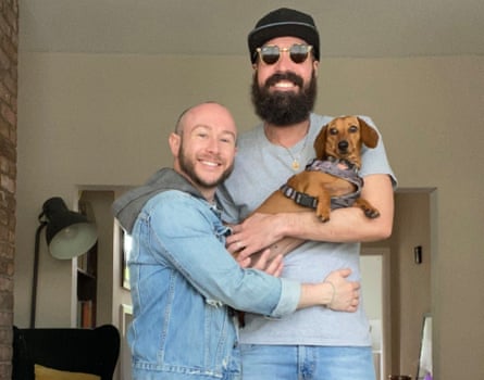 A man with shaved head with his arms around his partner who has a beard and sunglasses and is holding a smooth-haired dachshund