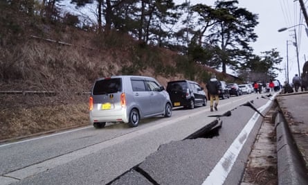 People walk past parked cars at the side of a road damaged by the earthquake