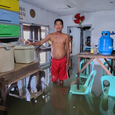 A man with no top on stands in a flooded room with plastic chairs and boxes and a gas canister on tables