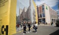 A huge computer-generated image on plastic sheeting covering a construction site shows a pleasant, modern shopping street. Pedestrians walk past the mural.