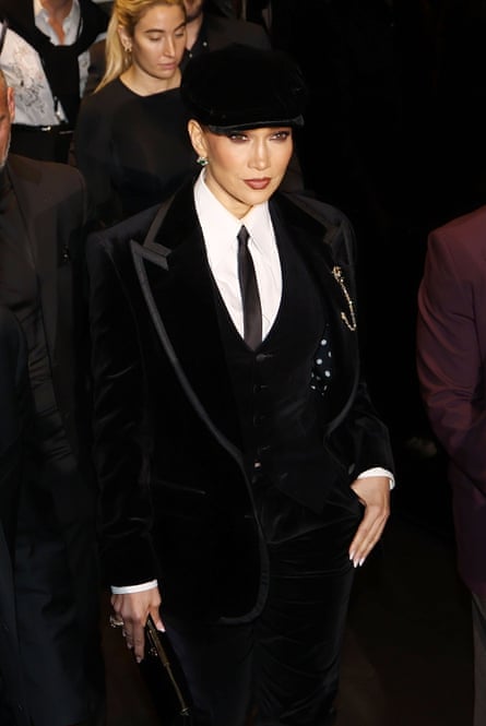 A woman, actor and singer Jennifer Lopez, in a black suit and tie, wearing a black hat, stands in a fashionable pose.