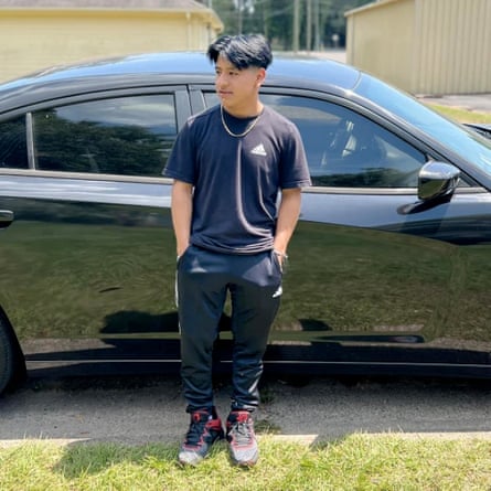 A Latino teenage boy, smiling, with thick black hair parted in the middle, wearing a black Adidas T-shirt and pants and red and black sneakers, and a hain, hands in pockets, standing beside a black car, smiling and looking to the right.