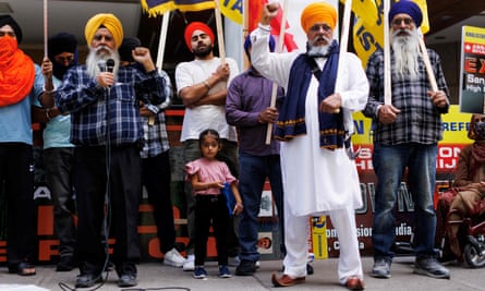 Group of mostly Indian men wearing orange and red turbans, and one little girl in a pink T-shirt, holding signs.