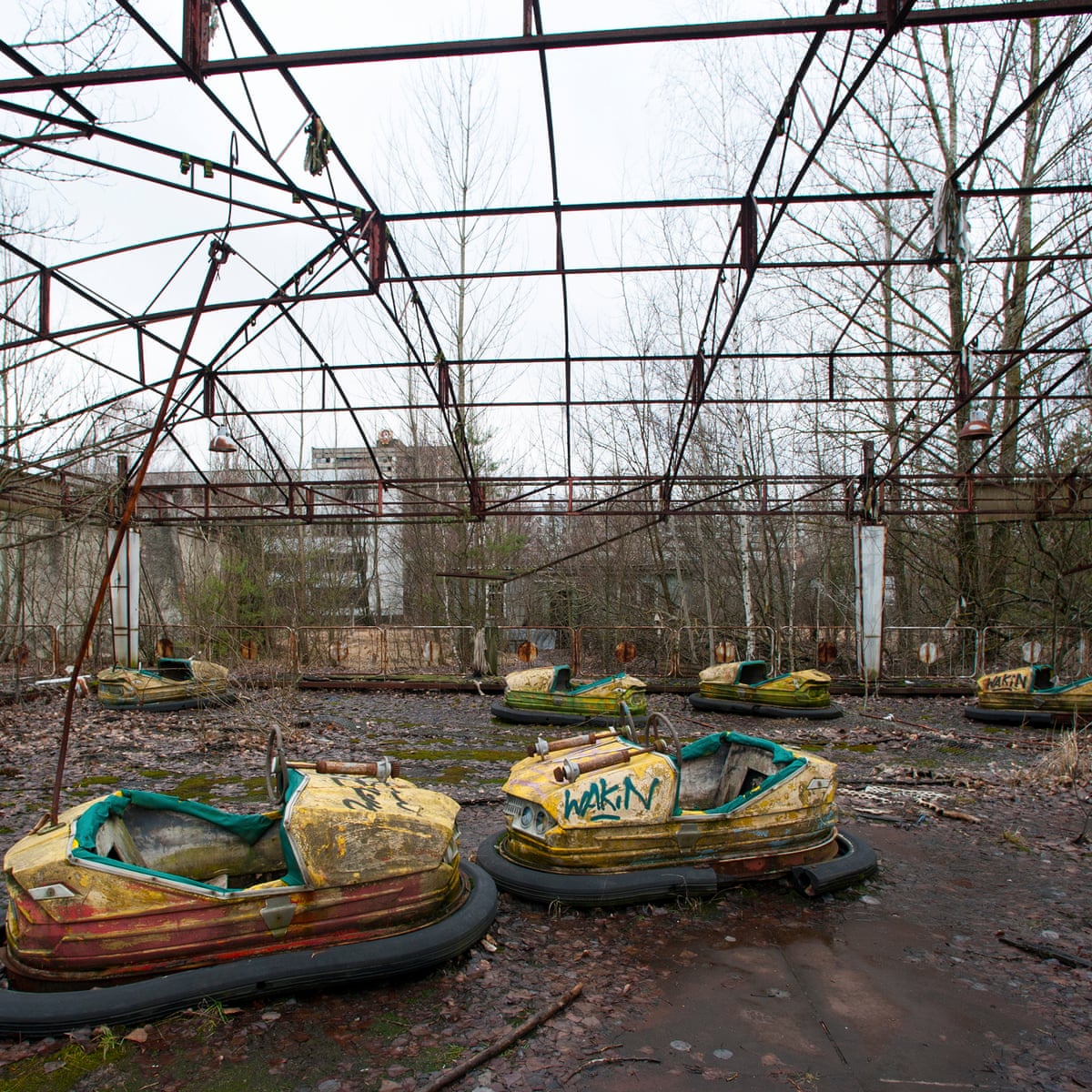 Abandoned bumper cars with graffiti are seen in a derelict amusement park within the deserted city of Chernobyl, surrounded by leafless trees and a collapsed metal frame overhead.