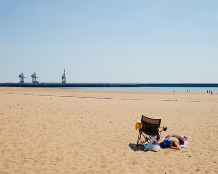 A woman sunbathing on a sandy beach with the works in the distance.